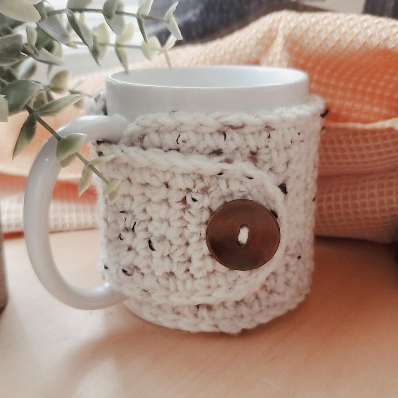 May include: White ceramic mug with a crocheted cozy in a cream color with a brown button. The cozy has a textured look and is wrapped around the mug.