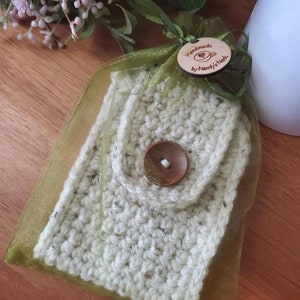 May include: A crocheted white and green cup cozy with a wooden button. The cozy is packaged in a green organza bag with a wooden tag that reads "Handmade by Nancy's Nook".