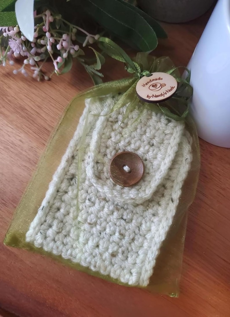 May include: A crocheted white and green cup cozy with a wooden button. The cozy is packaged in a green organza bag with a wooden tag that reads "Handmade by Nancy's Nook".