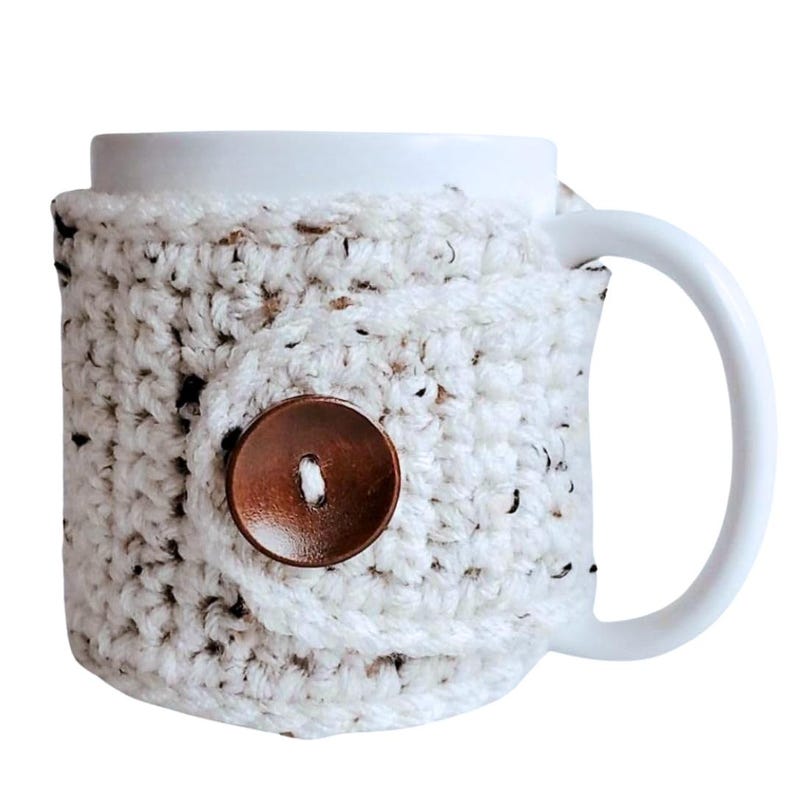 May include: White ceramic mug with a crocheted cozy in a speckled white and brown pattern. The cozy has a brown button.