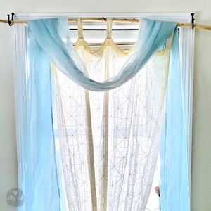 May include: A window with a light blue sheer curtain draped over a white sheer curtain with a geometric pattern. The curtains are hung on a natural wood rod.