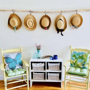 May include: A white shelf unit with two wicker baskets on each shelf. The shelf unit is in front of two yellow chairs with green cushions. Each chair has a pillow with a blue butterfly design. There are five straw hats hanging on a wooden rod above the shelf unit.