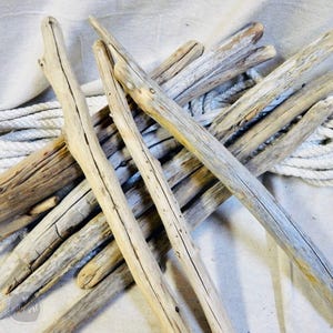 May include: A collection of weathered driftwood pieces in varying lengths and shades of gray and brown. The natural wood sticks have visible cracks and textures, with a white rope in the background. Ideal for crafting or coastal decor.