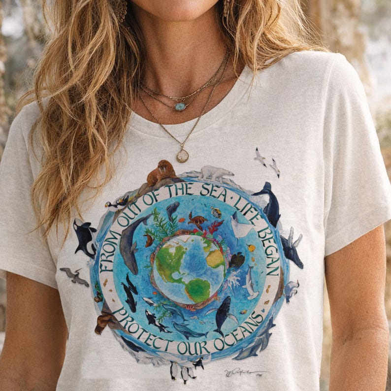 A woman wearing a white t-shirt with a graphic design depicting a globe surrounded by various marine animals, including penguins, and the text "FROM OUT OF THE SEA - LIFE BEGINS - PROTECT OUR OCEANS".