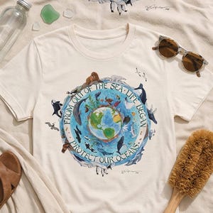 A white t-shirt with a graphic design depicting a globe surrounded by various marine life, along with the text "FROM OUT OF THE SEA LIFE BEGINS PROTECT OUR OCEANS".