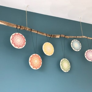May include: Six colorful string art mandalas hanging from a branch. Each mandala is a different color and has a unique design.