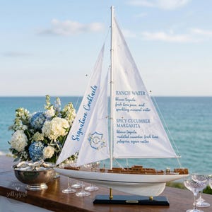 May include: A white model sailboat with two sails featuring blue text that reads Signature Cocktails, Ranch Water, and Spicy Cucumber Margarita. The boat has a wooden deck and hull, displayed on a black stand with a blue ocean background.