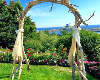May include: A rustic wooden archway, crafted from branches, stands on a green lawn. Cream-colored fabric drapes from the arch, with a backdrop of lush greenery, colorful flowers, and a distant ocean view under a bright blue sky. A lighthouse is visible in the distance.