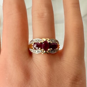 May include: A gold ring featuring three oval-cut deep red rubies set horizontally in the center, flanked by two curved bands encrusted with small, clear, round-cut white diamonds. The metal band has a polished yellow gold finish.