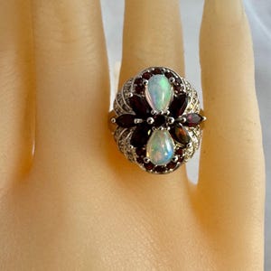 May include: A silver ring featuring a floral design with two pear-shaped opals and dark red garnet stones. The ring is adorned with small, clear accent stones around the opals and garnets. The ring is on a light skin tone.