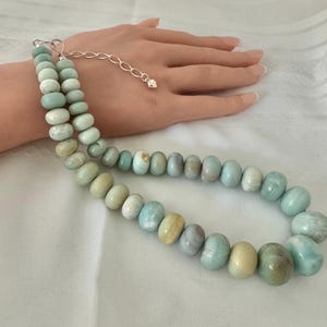 May include: A necklace featuring smooth, round, pale blue and green gemstone beads strung together. The piece includes a silver-toned metal chain extender with a small heart-shaped charm at the end, allowing for adjustable length and secure closure.