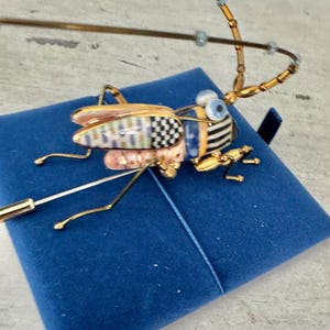 May include: A decorative insect pin with gold-tone metal legs, antennae, and wings. The insect's body features a mix of colors and patterns, including black and white checkerboard, stripes, and pink and blue accents. The pin is set on a blue velvet surface.