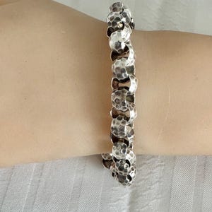 May include: A silver-toned bracelet with a hammered texture. The bracelet is composed of interconnected, rounded links. The links have a dimpled surface, creating a textured appearance. The bracelet is worn on a light-toned arm.