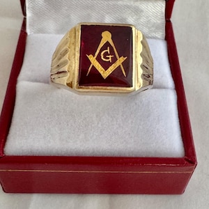 May include: A gold-toned ring with a rectangular, burgundy-colored center. The center features a gold emblem of a compass and square. The ring is presented in a red jewelry box with a white interior.
