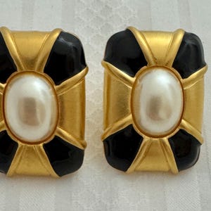May include: A pair of gold tone earrings with black enamel and a faux pearl center. The earrings are square shaped with a cross design.