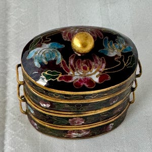 May include: A three-tiered, oval-shaped, black enamel box with a gold lid. The lid is decorated with floral designs in shades of blue, red, white, and green. The box is decorated with a similar floral design.