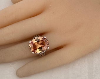 May include: A silver ring featuring a large, oval-cut peach-colored gemstone. The gemstone is the focal point, set in a silver band with a detailed design. The ring is displayed on a hand.