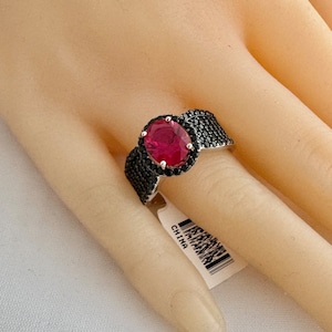 May include: A silver ring with a large red gemstone surrounded by a halo of smaller black gemstones. The ring is on a mannequin hand.