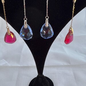 May include: Three pairs of dangle earrings with crystal drops. One pair has blue crystal drops, one pair has red crystal drops, and one pair has red crystal drops with a gold chain.