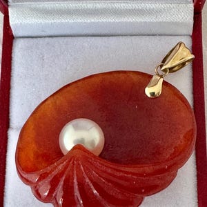 May include: A pendant featuring a carved, reddish-orange shell design with a white pearl at its center. The pendant has a gold-colored bail for hanging. The jewelry is presented in a red box with a white interior.