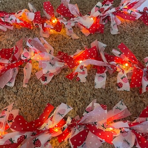 May include: A string of fairy lights wrapped in pink and red fabric with a strawberry print. The lights are strung together with a white cord.