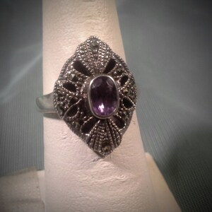 May include: A silver ring with a purple amethyst gemstone set in a filigree design with marcasite accents.