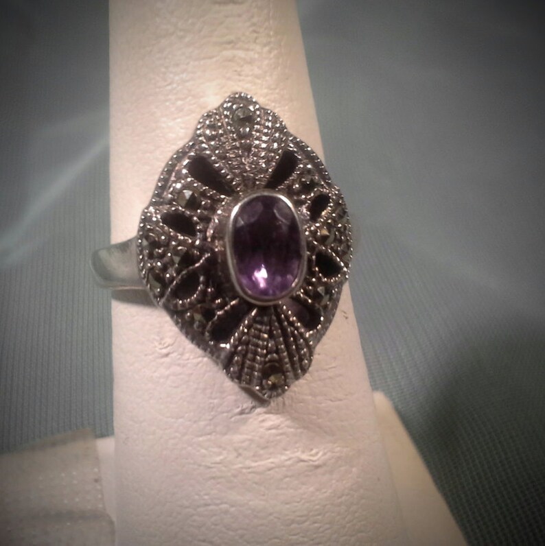 May include: A silver ring with a purple amethyst gemstone set in a filigree design with marcasite accents.