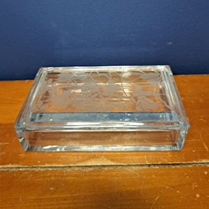 May include: A clear glass box with an etched floral design on the lid. The box is sitting on a wooden surface.
