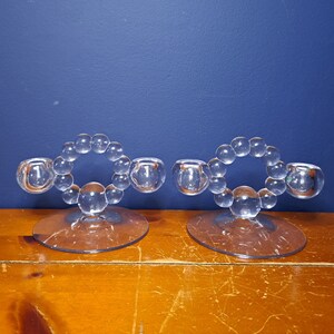 May include: Two clear glass candle holders with a beaded design. The holders are on a wooden surface.