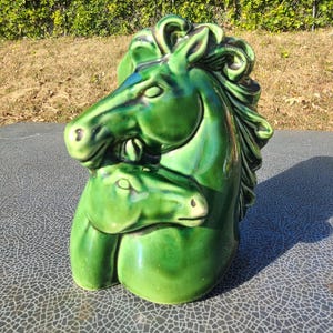 May include: A glossy, green ceramic sculpture of two horses, one embracing the other. The sculpture has a smooth, reflective surface and is set against a backdrop of green foliage and a blue sky. The horses' manes and features are detailed.