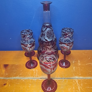 May include: A ruby red glass decanter with a matching set of four wine glasses. The decanter and glasses feature intricate etched floral designs. The set is displayed on a wooden surface against a blue background.