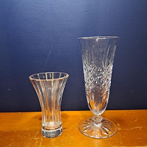 Puede incluir: Dos jarrones de cristal transparente sobre una superficie de madera. El jarr&oacute;n m&aacute;s alto tiene un patr&oacute;n de diamantes, mientras que el m&aacute;s corto tiene l&iacute;neas verticales. Ambos jarrones est&aacute;n vac&iacute;os y parecen ser decorativos.