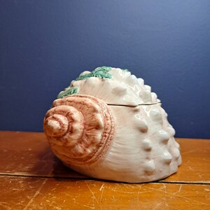 May include: A ceramic seashell-shaped container with a pink and brown glaze. The lid is also shaped like a seashell and has green leaves sculpted on top.