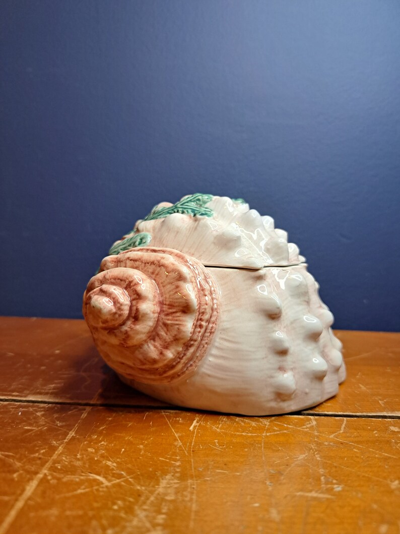 May include: A ceramic seashell-shaped container with a pink and brown glaze. The lid is also shaped like a seashell and has green leaves sculpted on top.