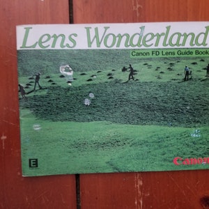 Könnte beinhalten: Ein grün-weißes Heft mit dem Titel "Lens Wonderland" und dem Untertitel "Canon FD Lens Guide Book". Das Cover zeigt ein Foto eines Golfplatzes mit Personen, die Golf spielen.