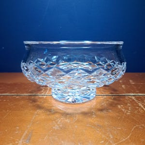 Może przedstawiać: Przezroczysta, szlifowana szklana miska z diamentowym wzorem wokół podstawy i gładką krawędzią. Misa stoi na krótkim, okrągłym cokole. Tło jest jednolicie niebieskie.