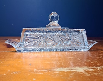 Op de afbeelding: Een heldere glazen botervloot met een deksel. De schaal heeft een rechthoekige basis en een deksel met een sterrenpatroon. Het deksel is bekroond met een facetgeslepen, diamantvormige handgreep. De schaal staat op een houten oppervlak.