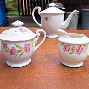May include: A white tea set with pink floral designs. The set includes a teapot, creamer, and sugar bowl with lid.