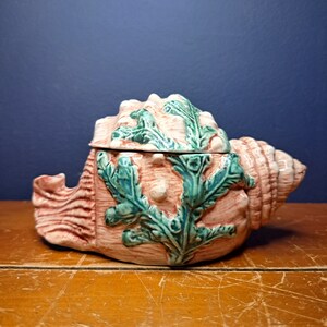 May include: A ceramic seashell shaped sugar bowl with a lid. The bowl is pink with green seaweed details.