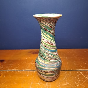 May include: A decorative vase with a flared top and a slender neck. The vase features a colorful, swirled pattern of green, brown, blue, and yellow. The vase is set against a blue background and a wooden surface.