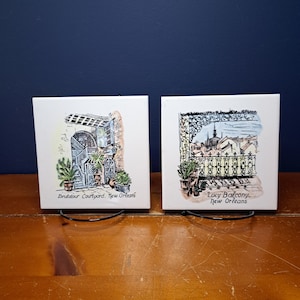 May include: Two white ceramic tiles with black metal stands. The tiles feature hand-drawn illustrations of New Orleans architecture. The first tile shows a courtyard with the text "Bridalour Courtyard, New Orleans". The second tile shows a balcony with the text "Lacy Balcony, New Orleans".