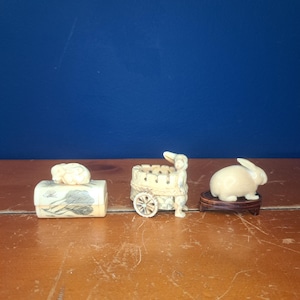 May include: Three small, off-white carved figurines of rabbits and a cart. One rabbit sits atop a rectangular box with a painted design. The cart has a rabbit on top and a wheel. The third rabbit sits on a small wooden stand.