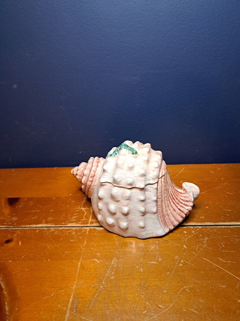 May include: A pink and white seashell shaped ceramic trinket box with green accents. The box is closed and sitting on a wooden surface.