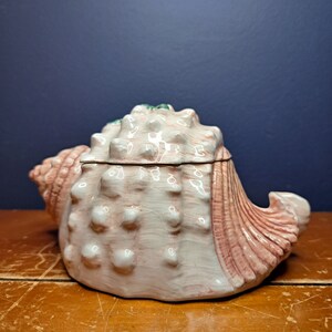 May include: A ceramic seashell trinket box with a pink and white glaze. The box is detailed with raised bumps and ridges, giving it a realistic seashell appearance.