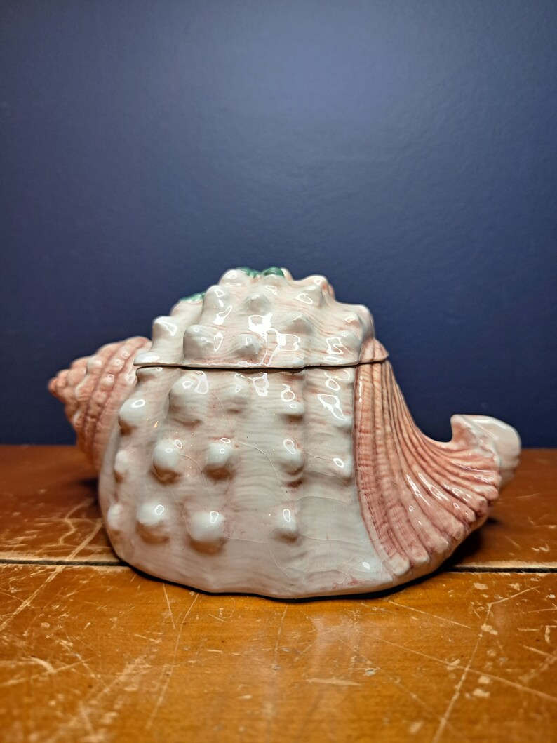 May include: A ceramic seashell trinket box with a pink and white glaze. The box is detailed with raised bumps and ridges, giving it a realistic seashell appearance.