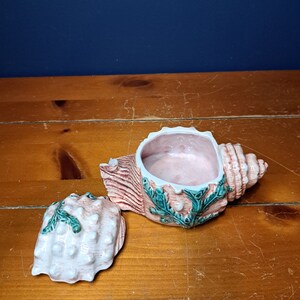 May include: A ceramic seashell trinket box with a pink and white shell design and green seaweed accents. The lid is open and the inside of the box is pink.