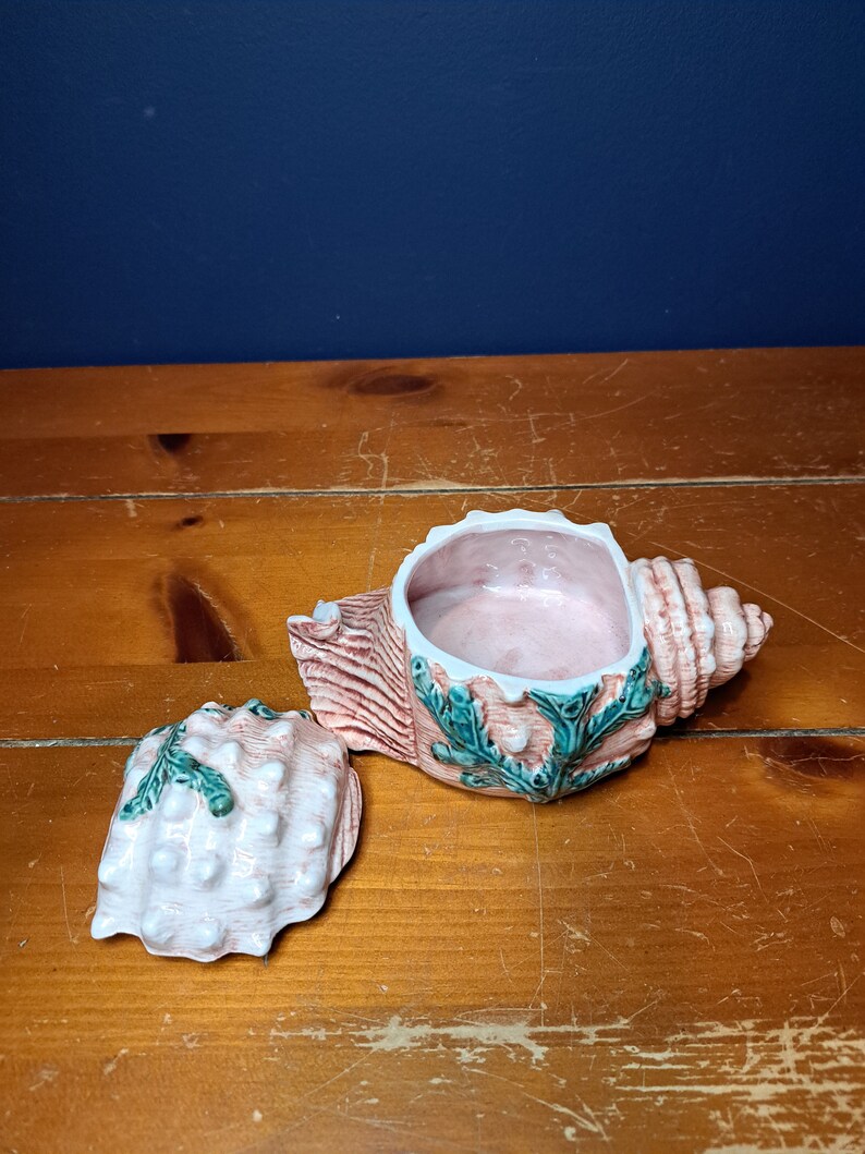 May include: A ceramic seashell trinket box with a pink and white shell design and green seaweed accents. The lid is open and the inside of the box is pink.