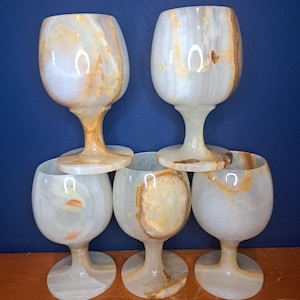 May include: A set of six onyx wine glasses. The glasses have a rounded bowl shape and a stem. The onyx is white with brown and tan veining. The glasses are arranged in a pyramid shape against a blue background.