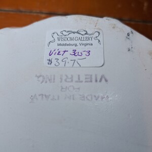 May include: A white ceramic plate with a sticker that reads "Wisdom Gallery, Middleburg, Virginia" and "Viet 3053 $39.75". The plate also has a sticker that says "Vietri Inc. For Made in Italy".
