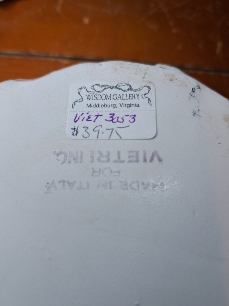May include: A white ceramic plate with a sticker that reads "Wisdom Gallery, Middleburg, Virginia" and "Viet 3053 $39.75". The plate also has a sticker that says "Vietri Inc. For Made in Italy".